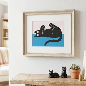 May include: Framed artwork featuring a black cat in a playful pose, set against a pink and blue pixelated background. The artwork is displayed on a light-colored wooden shelf, alongside two black cat figurines and a potted succulent.