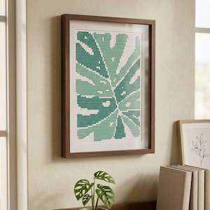 Pode incluir: Uma obra de arte em ponto cruz emoldurada com um grande design de folha de monstera verde. A obra de arte é exibida em uma moldura de madeira marrom escura e pendurada em uma parede bege. Uma pequena planta em vaso e livros estão em uma prateleira de madeira abaixo.