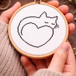 Puede incluir: Un bastidor de bordado con un diseño de gato negro dentro de un corazón. El gato está durmiendo y el diseño está sobre tela blanca. El bastidor es de madera y está sostenido por manos.