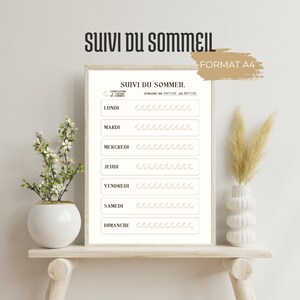 Peut inclure: Un tableau de suivi du sommeil encadré, intitulé "SUIVI DU SOMMEIL" et portant la mention "FORMAT A4". Le tableau blanc présente du texte noir, les jours de la semaine et des icônes de lune. Il est posé sur une étagère en bois avec des vases.