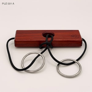 May include: A wooden puzzle with a dark reddish-brown rectangular block, a black rope, and two silver metal rings. The block has a hole in the center and the text "PUZ 001 A" is visible in the image.