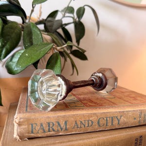 May include: A vintage door knob set featuring two clear, faceted glass knobs connected by a central metal spindle. The glass knobs have an octagonal shape with a reflective surface, while the connecting rod is made of dark, weathered metal.