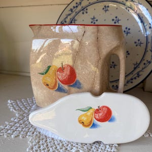 May include: A beige ceramic pitcher with a red rim and a visible crack, featuring a painted pear and apple design. In front lies a matching white ceramic lid with an identical pear and apple illustration, both displaying a glossy finish.