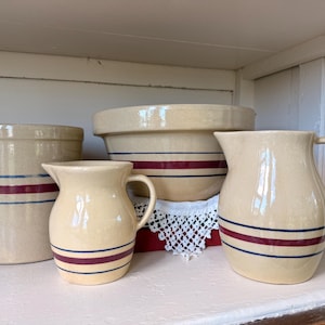 May include: A collection of cream-colored stoneware featuring a cylindrical crock, a large mixing bowl, and two pitchers. Each piece is decorated with horizontal bands in dark blue and deep red stripes, creating a uniform set of kitchenware.