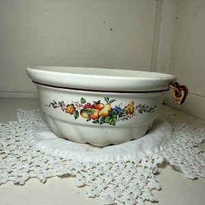 May include: Cream-colored ceramic bowl with a fruit and floral design around the rim. The bowl has a brown rim and a decorative handle. It sits on a white, crocheted doily.