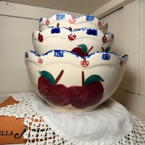 May include: A stack of three ceramic bowls with a cream-colored base. Each bowl features a scalloped blue and white checkered rim and hand-painted red apples with green leaves. The bowls are nested on a white doily.