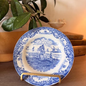 May include: A round ceramic plate featuring a blue and white transferware pattern. The center depicts a coastal scene with figures, sailboats, and a windmill. The rim is decorated with an intricate floral and scroll border design.