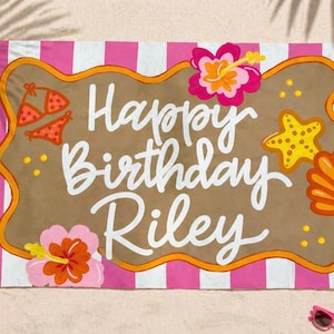 Może przedstawiać: Ręcznik plażowy w różowo-białe paski. Na brązowym środku napis "Happy Birthday Riley" białym pismem. Ozdobiony bikini, rozgwiazdami i kwiatami, idealny na urodziny na plaży.