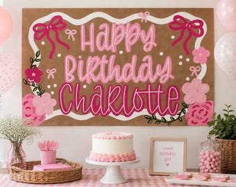 May include: A rectangular brown sign with white wavy borders, pink bows, and floral accents, featuring the text Happy Birthday Charlotte in pink and dark pink. Below, a white cake with pink frosting sits on a stand near a framed sign.