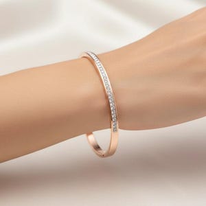 May include: Rose gold bangle bracelet with a row of clear crystals. The bracelet is on a person's arm, showcasing the jewelry's design and sparkle. The background is a soft, neutral color.