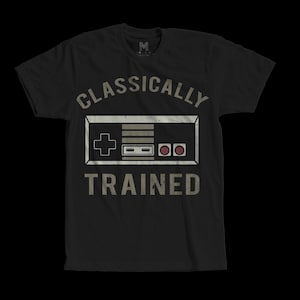 Könnte beinhalten: Schwarzes T-Shirt mit einem Retro-Videospiel-Controller-Design. Der Text "CLASSICALLY TRAINED" ist oberhalb und unterhalb des Controllers in einer verblichenen Schriftart aufgedruckt. Das T-Shirt hat einen Rundhalsausschnitt und kurze Ärmel.