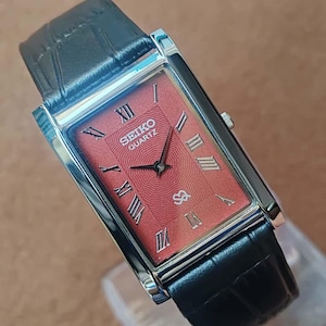 May include: A rectangular Seiko Quartz watch with a red face and black leather strap. The watch face features Roman numerals and the Seiko logo. The watch has a silver bezel and a black leather strap with a crocodile pattern.