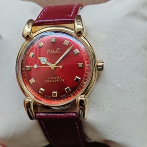 May include: A gold-toned Piaget wristwatch with a vibrant red face and a matching burgundy leather strap. The watch features gold hands and markers, and the face displays the brand name and "17 Jewels Shock Proof" text. The watch is displayed on a white surface.