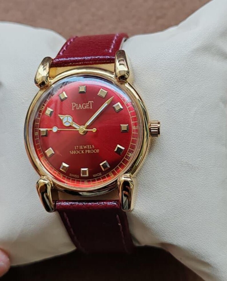 May include: A gold-toned Piaget wristwatch with a vibrant red face and a matching burgundy leather strap. The watch features gold hands and markers, and the face displays the brand name and "17 Jewels Shock Proof" text. The watch is displayed on a white surface.