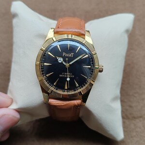 May include: A gold-toned Piaget wristwatch with a black face and a brown leather band. The watch face features gold hands and markers, with the brand name and "17 Jewels Shock Proof" text. The watch is displayed on a cream-colored pillow.