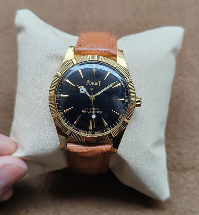 May include: A gold-toned Piaget wristwatch with a black face and a brown leather band. The watch face features gold hands and markers, with the brand name and "17 Jewels Shock Proof" text. The watch is displayed on a cream-colored pillow.