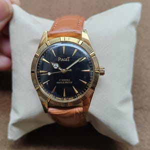 May include: A gold-toned Piaget watch with a black face and a brown leather strap. The watch face features gold hands and markers, with the text "Piaget" and "17 Jewels Shock Proof". The watch is displayed on a cream-colored pillow.
