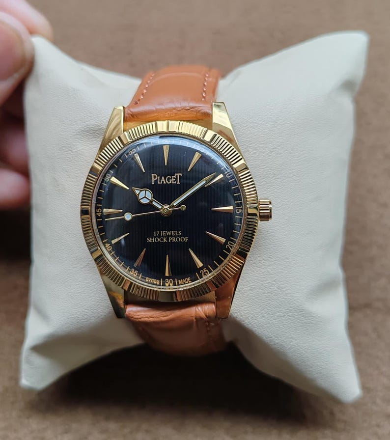 May include: A gold-toned Piaget watch with a black face and a brown leather strap. The watch face features gold hands and markers, with the text "Piaget" and "17 Jewels Shock Proof". The watch is displayed on a cream-colored pillow.
