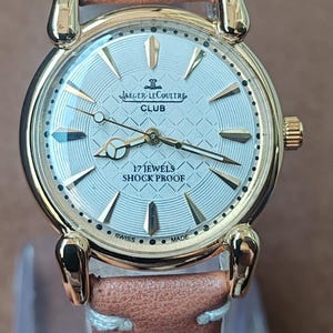 May include: A gold-toned Jaeger-LeCoultre Club watch with a light-colored face and a brown leather band. The watch face features gold hands and markers, with the text "17 Jewels Shock Proof" and "Swiss Made".