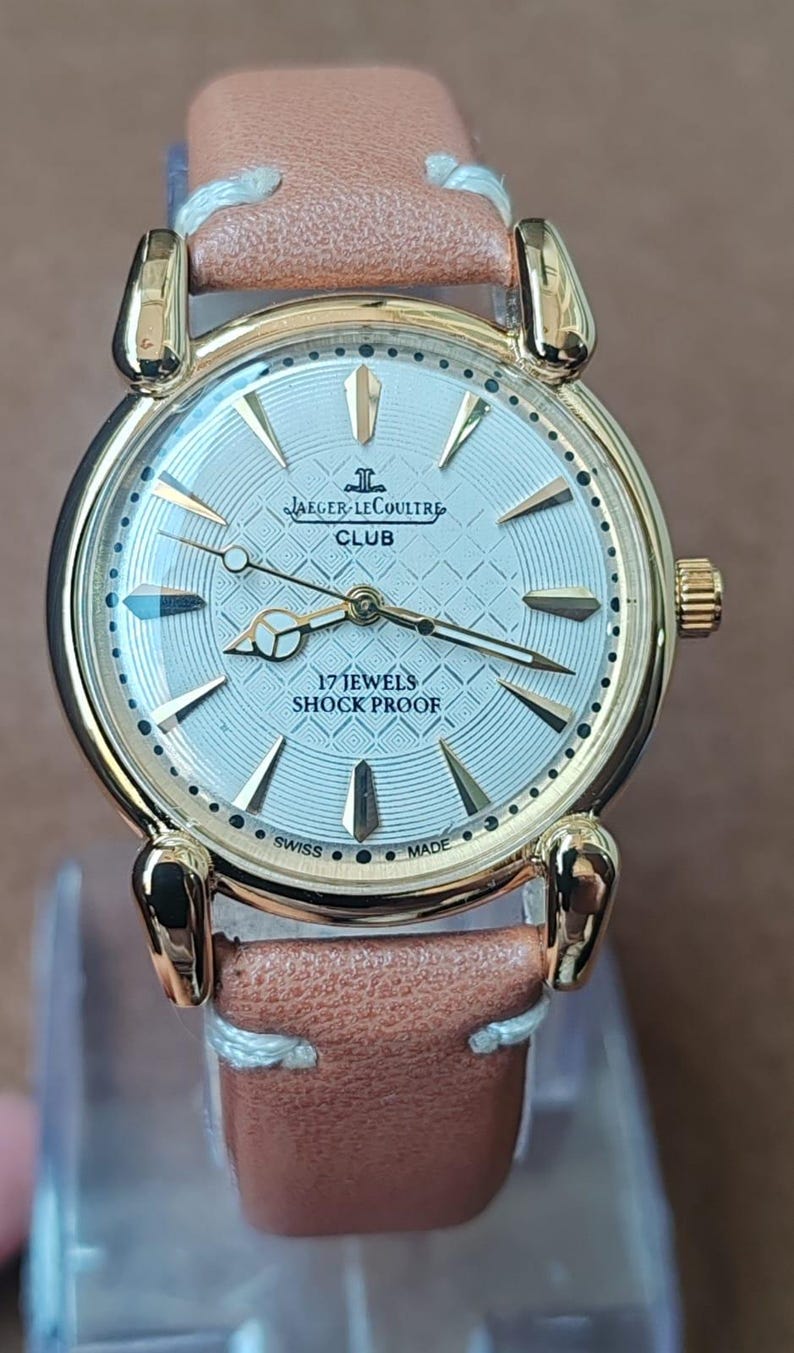 May include: A gold-toned Jaeger-LeCoultre Club watch with a light-colored face and a brown leather band. The watch face features gold hands and markers, with the text "17 Jewels Shock Proof" and "Swiss Made".