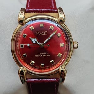 May include: A gold-toned Piaget watch with a deep red face and matching leather band. The watch face features gold markers and hands, with the text "Piaget", "17 Jewels Shock Proof", "Swiss Made".