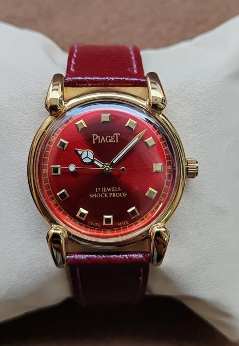 May include: A gold-toned Piaget watch with a deep red face and matching leather band. The watch face features gold markers and hands, with the text "Piaget", "17 Jewels Shock Proof", "Swiss Made".