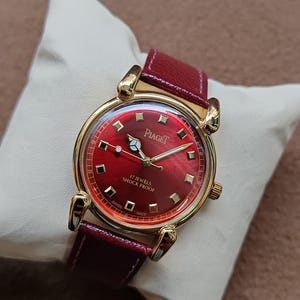May include: A vintage Piaget watch with a red dial and a gold-tone case. The watch features a red leather band and is displayed on a white pillow. The watch face has gold markers and hands. The text "Piaget" and "17 Jewels Shock Proof" are visible.