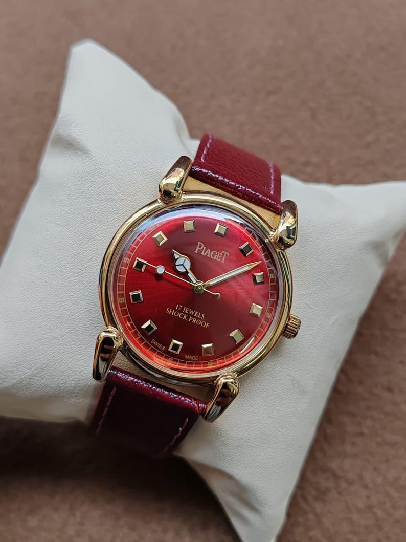 May include: A vintage Piaget watch with a red dial and a gold-tone case. The watch features a red leather band and is displayed on a white pillow. The watch face has gold markers and hands. The text "Piaget" and "17 Jewels Shock Proof" are visible.