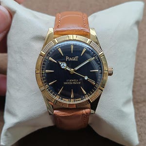 May include: A gold-toned Piaget wristwatch with a black face and a brown leather band. The watch face features gold hands and markers, with the text "Piaget" and "17 Jewels Shock Proof". The watch is displayed on a cream-colored pillow.