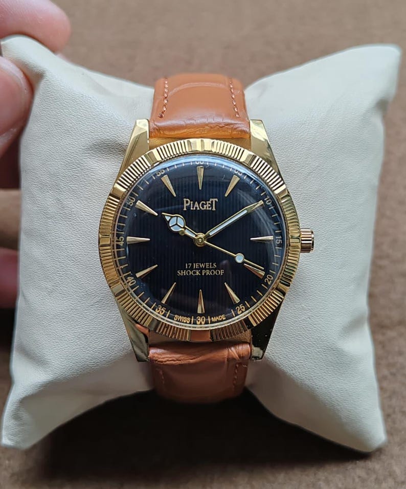 May include: A gold-toned Piaget wristwatch with a black face and a brown leather band. The watch face features gold hands and markers, with the text "Piaget" and "17 Jewels Shock Proof". The watch is displayed on a cream-colored pillow.