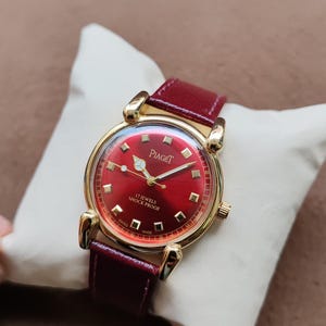 May include: A vintage Piaget watch with a red dial and a gold-tone case. The watch features a red leather band and is marked "17 Jewels Shock Proof" and "Swiss Made". The watch is displayed on a white pillow.