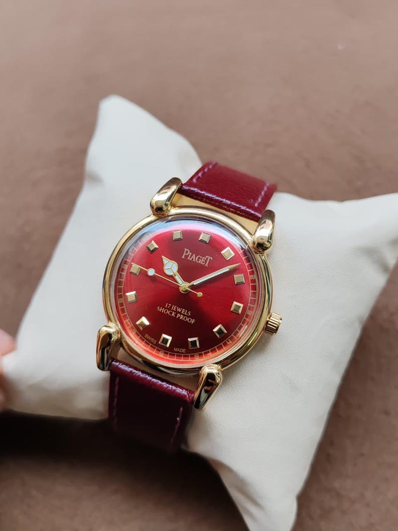 May include: A vintage Piaget watch with a red dial and a gold-tone case. The watch features a red leather band and is marked "17 Jewels Shock Proof" and "Swiss Made". The watch is displayed on a white pillow.
