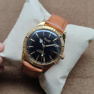 May include: A gold-toned Piaget watch with a black face and brown leather band. The watch face features gold hands and markers, with the brand name and "17 JEWELS SHOCK PROOF" text. The watch is displayed on a cream-colored pillow.