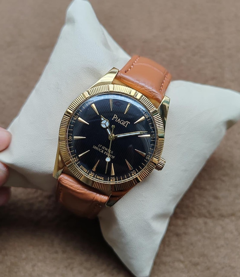 May include: A gold-toned Piaget watch with a black face and brown leather band. The watch face features gold hands and markers, with the brand name and "17 JEWELS SHOCK PROOF" text. The watch is displayed on a cream-colored pillow.