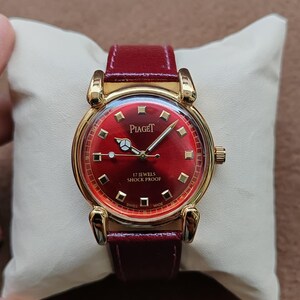 May include: A vintage Piaget watch with a red dial and a gold-tone case. The watch features a burgundy leather strap and the text "17 Jewels Shock Proof". The watch is displayed on a cream-colored pillow.