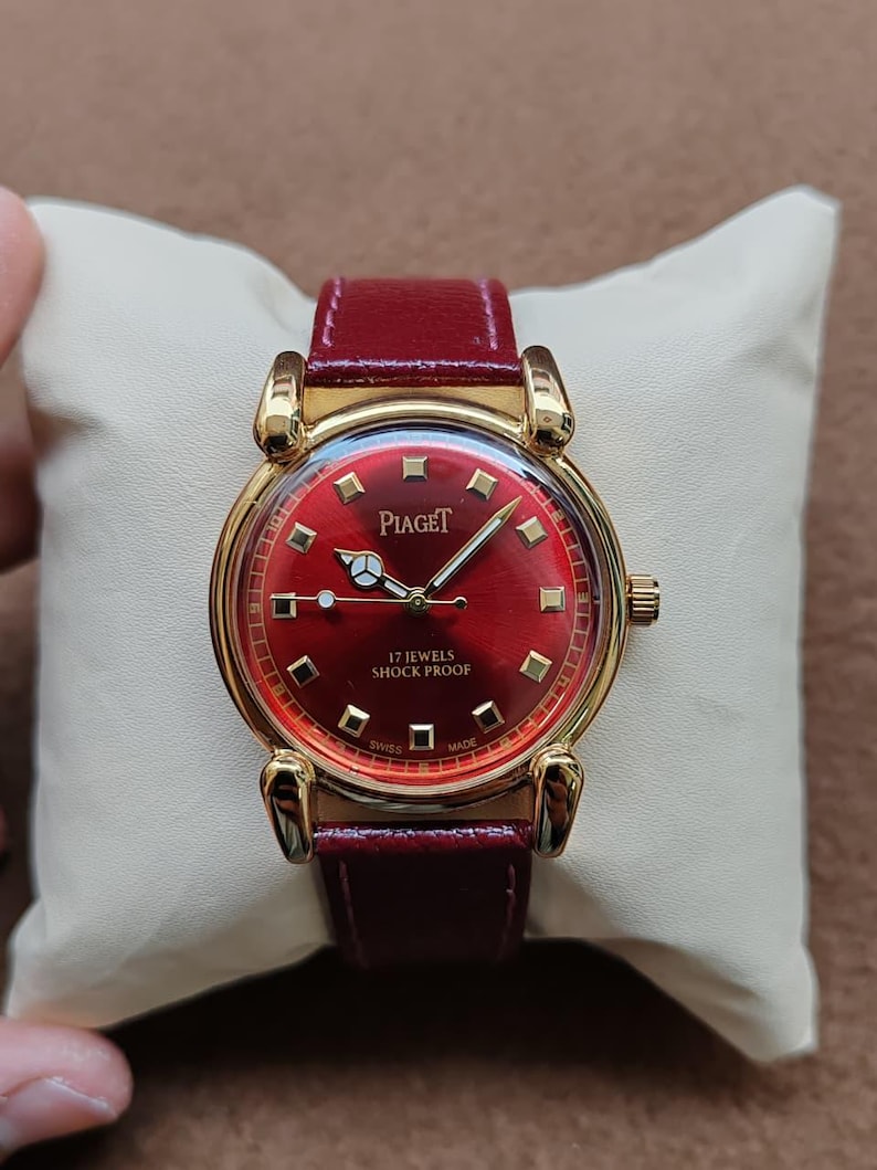 May include: A vintage Piaget watch with a red dial and a gold-tone case. The watch features a burgundy leather strap and the text "17 Jewels Shock Proof". The watch is displayed on a cream-colored pillow.
