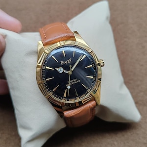 May include: A gold-toned Piaget wristwatch with a black face and a brown leather band. The watch face features gold hands and markers, with the brand name and "17 JEWELS SHOCK PROOF" printed on it. The watch is displayed on a cream-colored pillow.