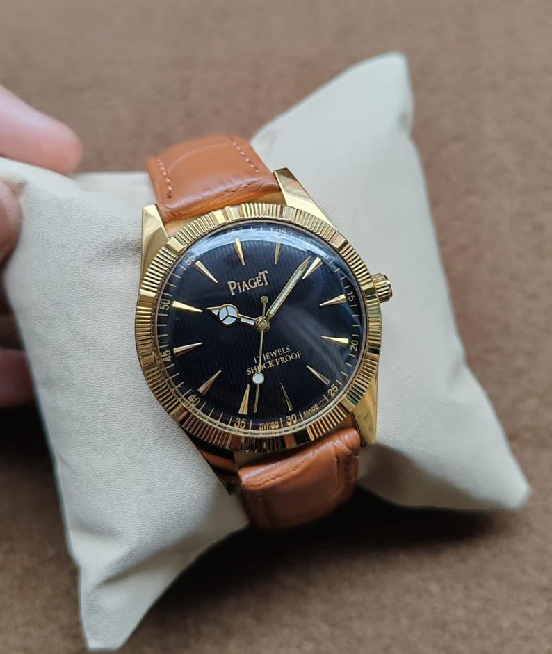 May include: A gold-toned Piaget wristwatch with a black face and a brown leather band. The watch face features gold hands and markers, with the brand name and "17 JEWELS SHOCK PROOF" printed on it. The watch is displayed on a cream-colored pillow.