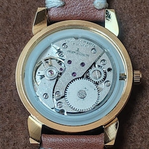 May include: A gold-toned wristwatch with a brown leather band. The watch face is transparent, revealing the intricate internal mechanisms, including gears and jewels. The band is secured with light-colored stitching.