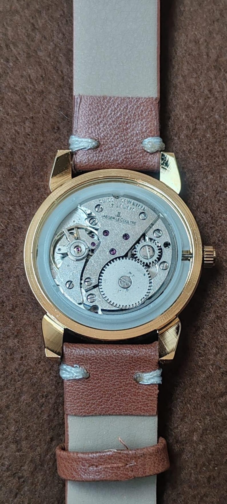 May include: A gold-toned wristwatch with a brown leather band. The watch face is transparent, revealing the intricate internal mechanisms, including gears and jewels. The band is secured with light-colored stitching.