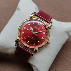 May include: A gold-toned Piaget wristwatch with a red dial and a red leather band. The watch face features gold hands and markers, with the brand name and "17 JEWELS SHOCK PROOF" printed on the dial. The watch is displayed on a white pillow.