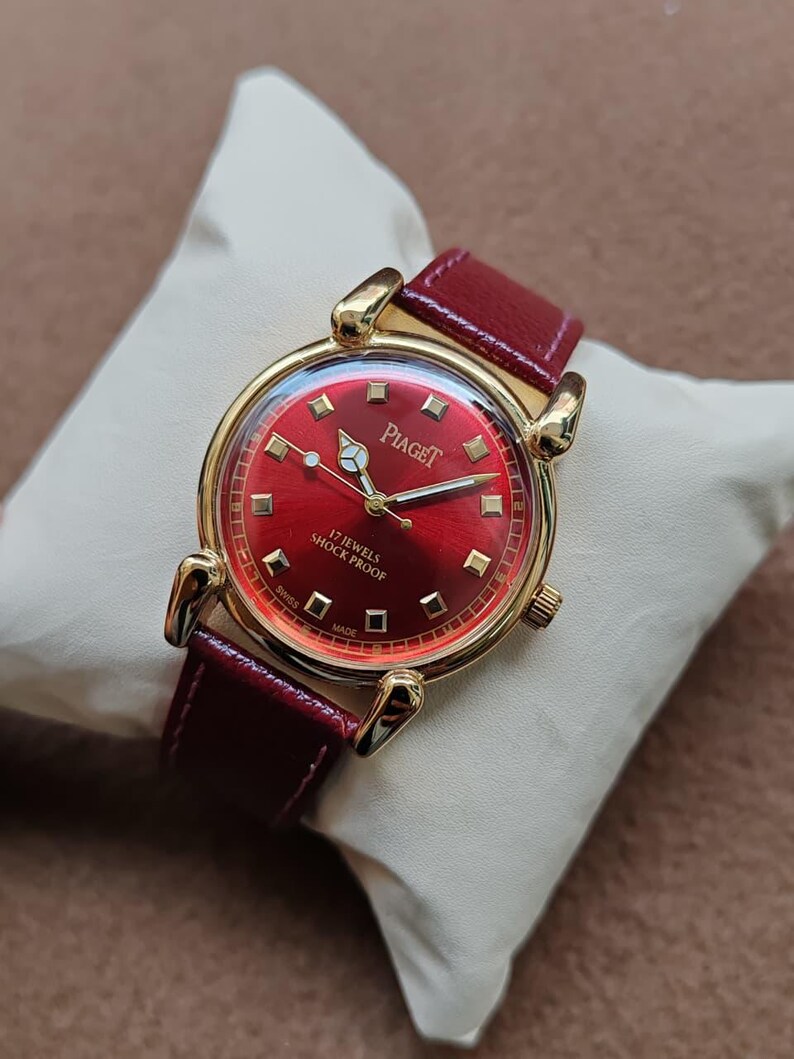 May include: A gold-toned Piaget wristwatch with a red dial and a red leather band. The watch face features gold hands and markers, with the brand name and "17 JEWELS SHOCK PROOF" printed on the dial. The watch is displayed on a white pillow.