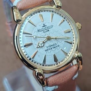 May include: A gold-toned Jaeger-LeCoultre Club watch with a light-colored face and a brown leather strap. The watch face features the brand name, the word "CLUB", and the text "17 JEWELS SHOCK PROOF". The watch is Swiss made.