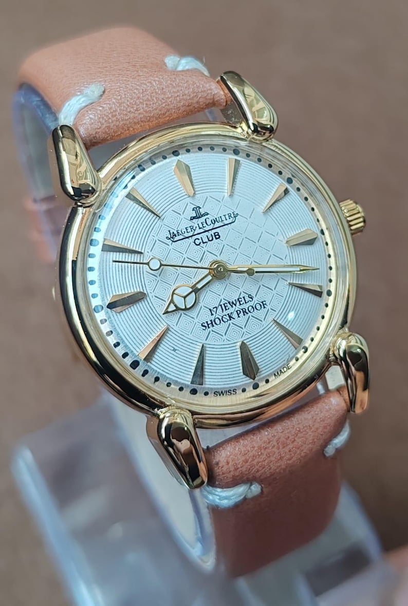 May include: A gold-toned Jaeger-LeCoultre Club watch with a light-colored face and a brown leather strap. The watch face features the brand name, the word "CLUB", and the text "17 JEWELS SHOCK PROOF". The watch is Swiss made.