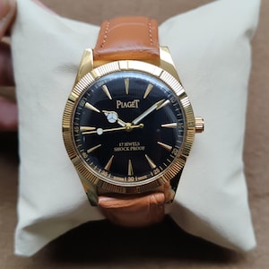 May include: A gold-toned Piaget wristwatch with a black face and a brown leather band. The watch face displays the brand name and the words "17 Jewels Shock Proof". The watch is set on a cream-colored cushion.