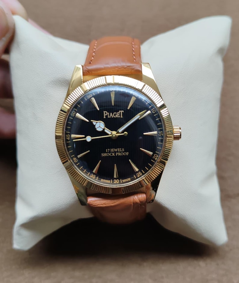 May include: A gold-toned Piaget wristwatch with a black face and a brown leather band. The watch face displays the brand name and the words "17 Jewels Shock Proof". The watch is set on a cream-colored cushion.