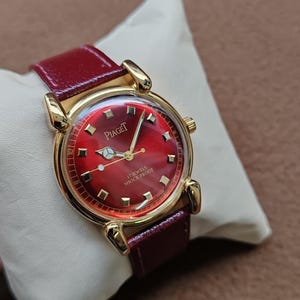 May include: A gold-toned Piaget wristwatch with a red face and a burgundy leather band. The watch face features gold-colored hour markers and hands. The text "Piaget" is visible on the watch face. The watch is labeled "17 Jewels Shock Proof".
