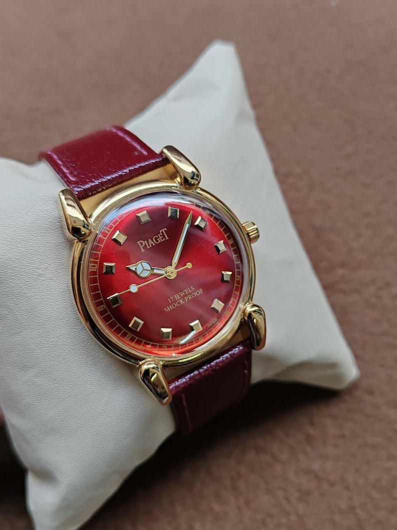 May include: A gold-toned Piaget wristwatch with a red face and a burgundy leather band. The watch face features gold-colored hour markers and hands. The text "Piaget" is visible on the watch face. The watch is labeled "17 Jewels Shock Proof".