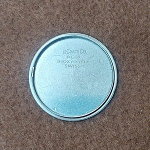 May include: A silver-colored watch component with the text "LE COULTRE CO ACER INOXIDABLE SWISS" engraved on its surface. The component is circular with a polished finish and is set against a brown background.