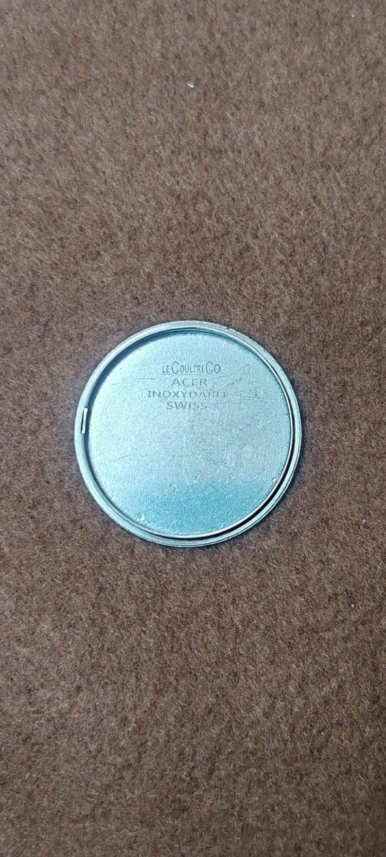 May include: A silver-colored watch component with the text "LE COULTRE CO ACER INOXIDABLE SWISS" engraved on its surface. The component is circular with a polished finish and is set against a brown background.